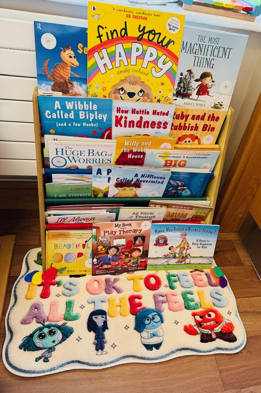 A shelf of children's picture books about feelings beside an 'It's OK to feel all the feels' rug